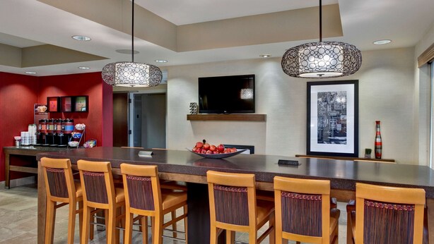 A modern kitchen with a large island, wooden chairs, a bowl of fruit, and a television above the island. - Hampton Inn West Des Moines Lake Drive (West Des Moines)