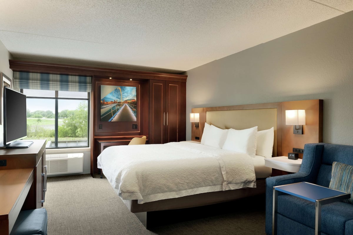 Ein Hotelzimmer mit einem großen Bett, einem Schreibtisch mit Computer, einem Sessel und einem Fenster mit Blick auf Grünflächen.