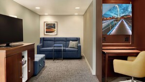 A hotel room with a blue sofa, a TV, a desk, and a large window with a cityscape view. - Hampton Inn West Des Moines Lake Drive (West Des Moines)