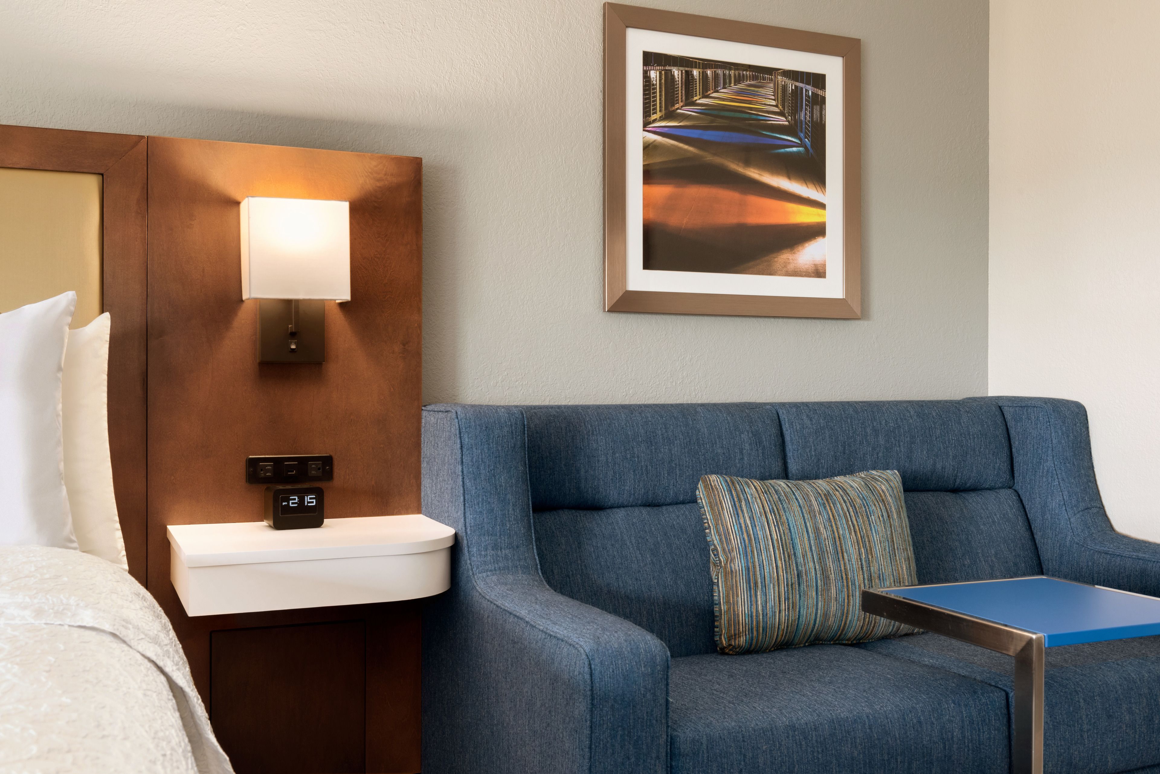 Habitación de hotel con sofá azul, una mesita de noche con lámpara, un cuadro en la pared y un reloj digital.