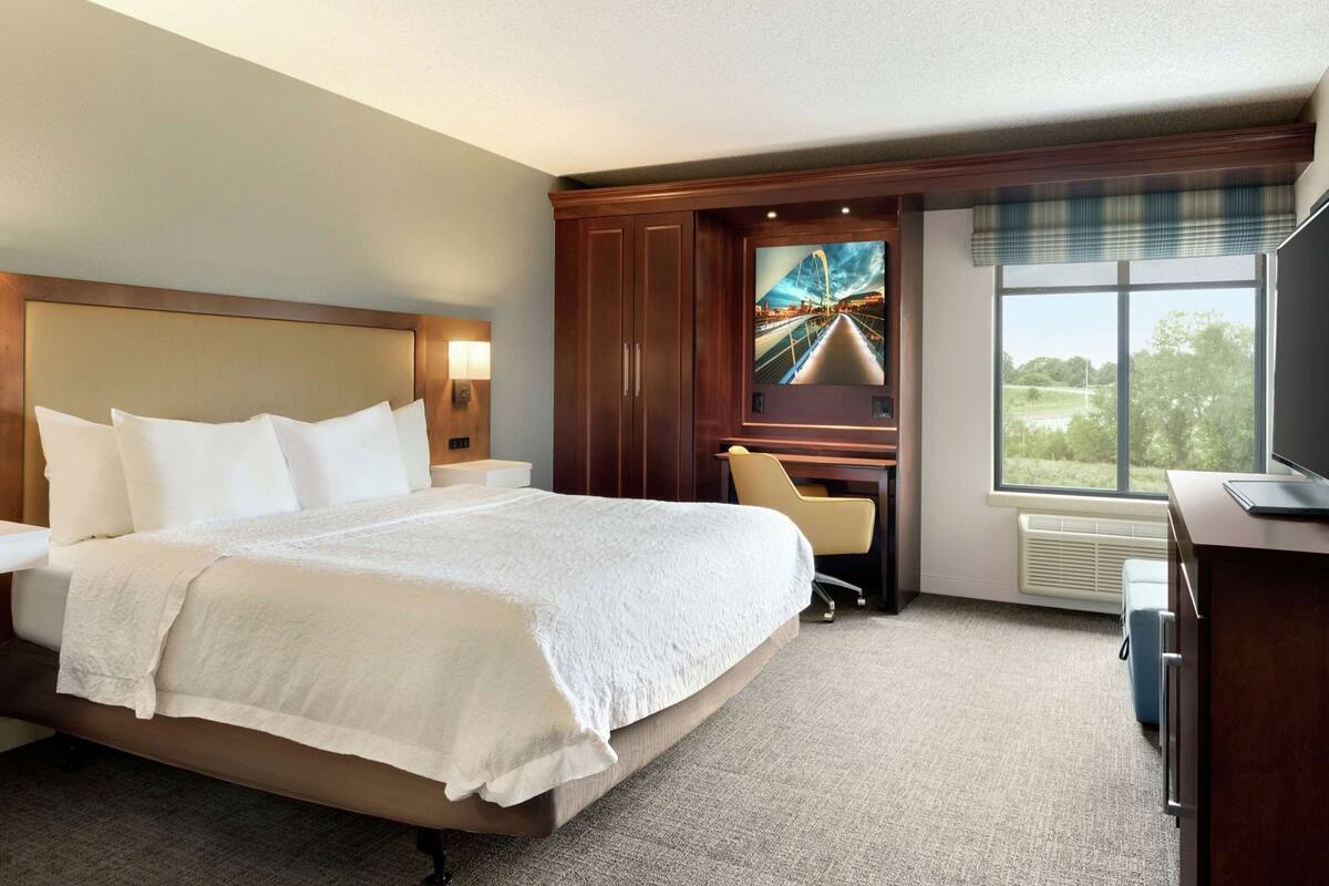 Ein Hotelzimmer mit einem großen Bett, einem Schreibtisch mit Stuhl, einem Fernseher und einem Fenster mit Blick auf Grünflächen.
