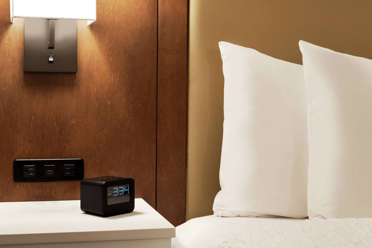 Ein Hotelzimmer mit Nachttisch, digitaler Uhr, Wandleuchte und einem Bett mit weißen Kissen.