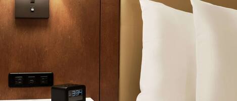 A hotel room with a bedside table, a digital clock, a wall-mounted light, and a bed with white pillows.