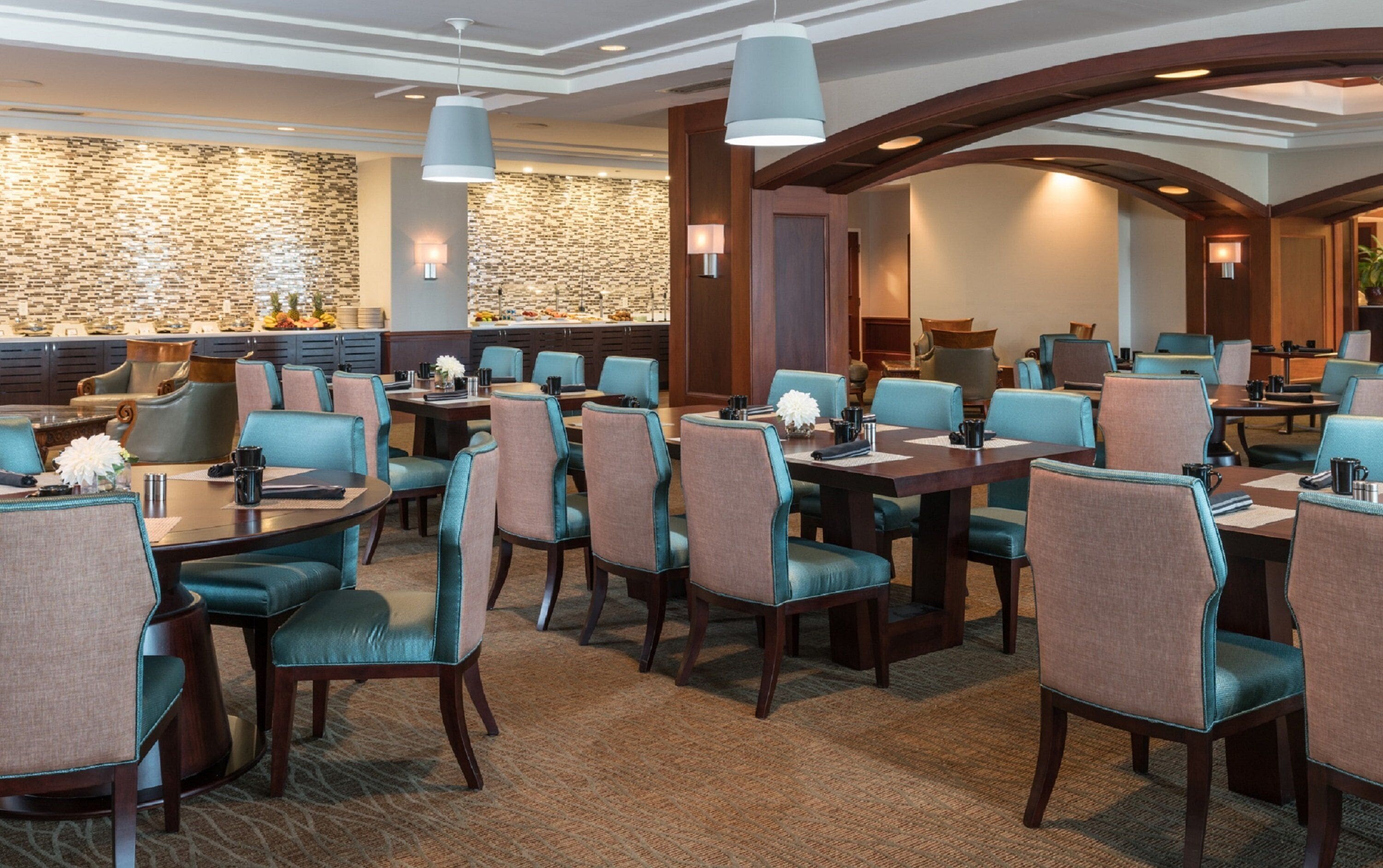 A hotel dining area with multiple tables and chairs, a stone wall, and modern lighting fixtures.