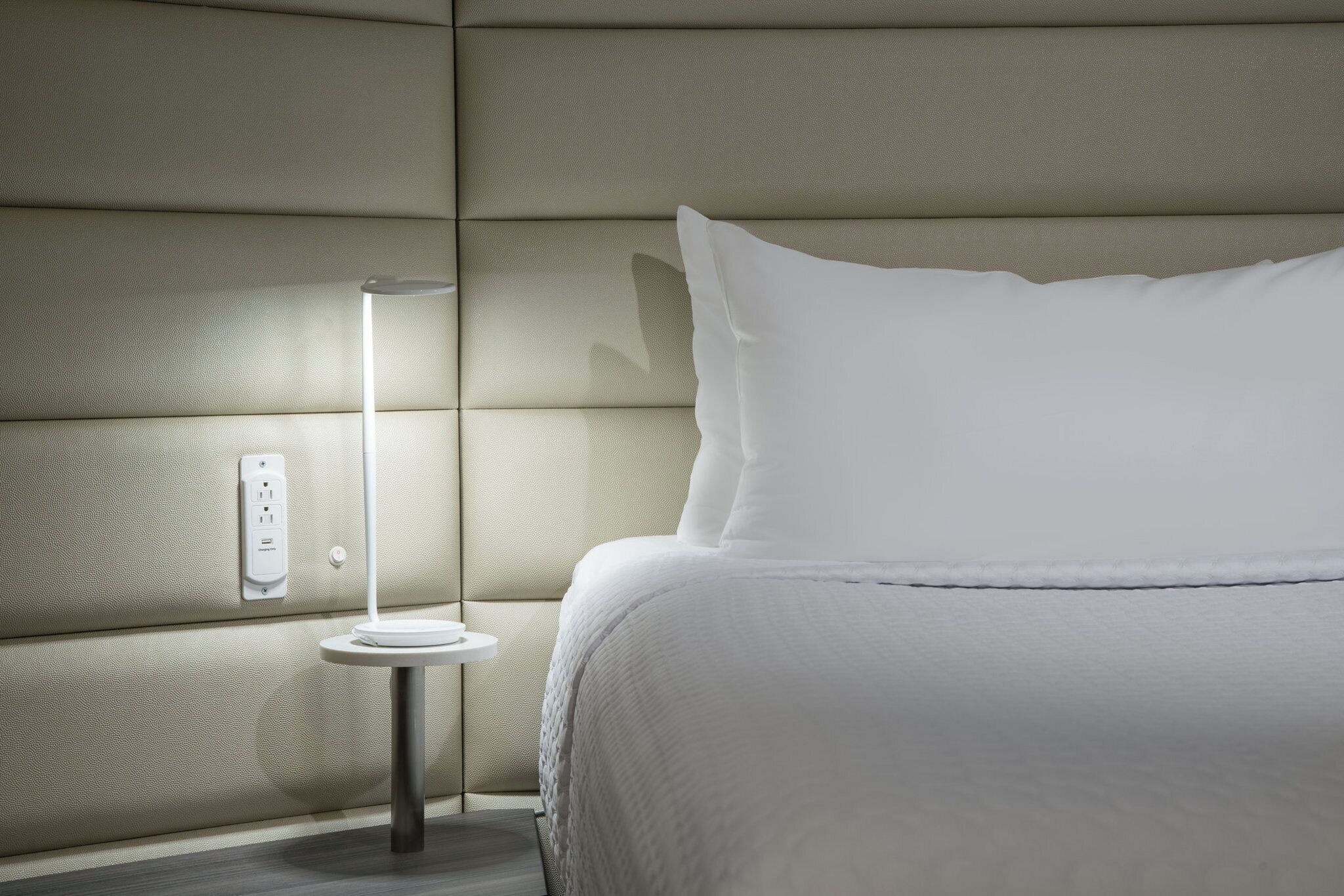A neatly made bed with white bedding, a bedside table with a lamp, and a wall socket.
