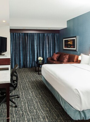 A hotel room with a bed, a desk with a chair, a television, a small table with a vase of flowers, and a painting on the wall.