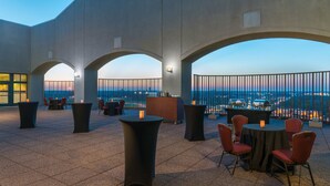 An outdoor terrace with tables and chairs, a bar area, and a view of the city skyline at dusk.