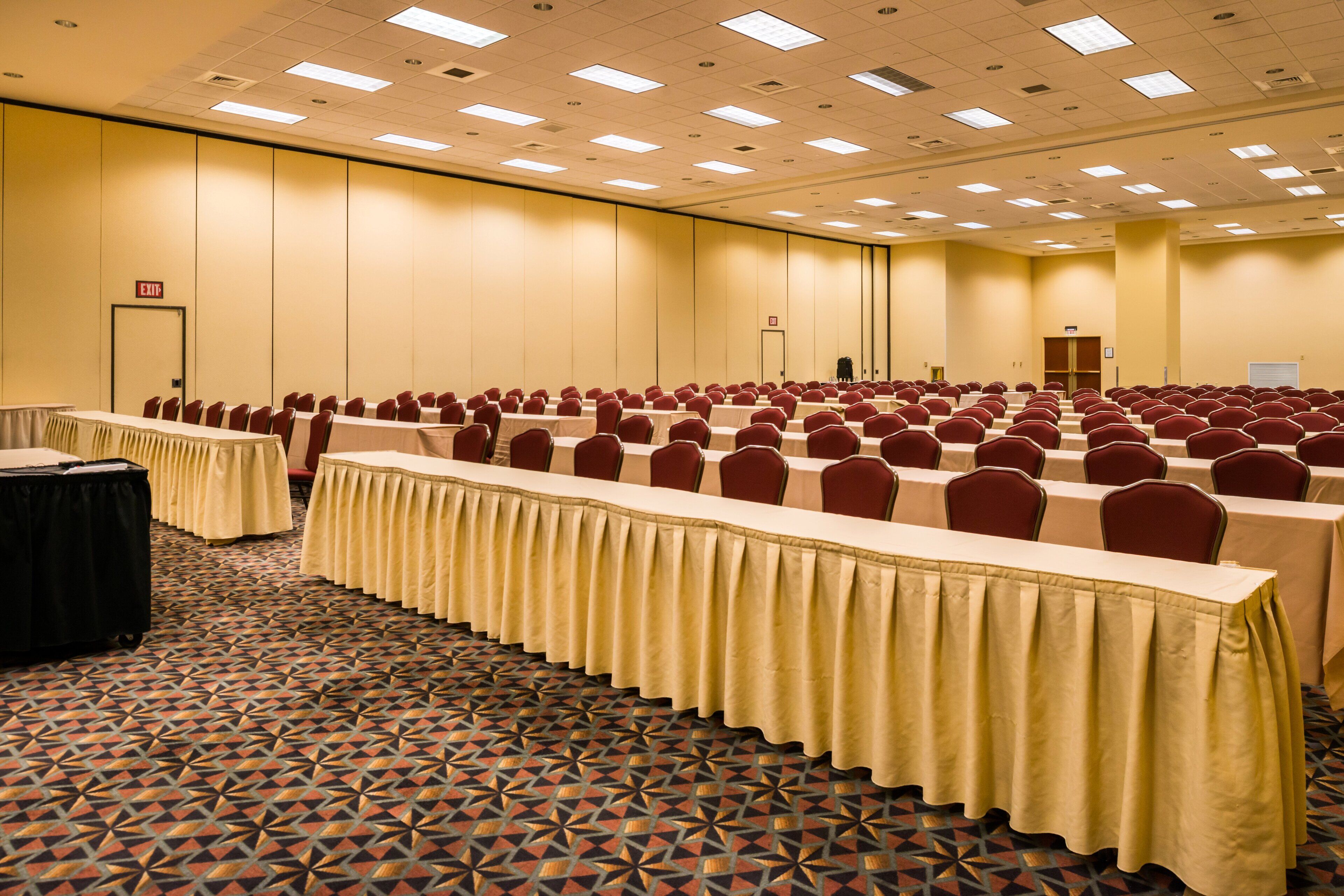 A conference room with rows of tables and chairs, carpeted floor, and a high ceiling with recessed lighting.