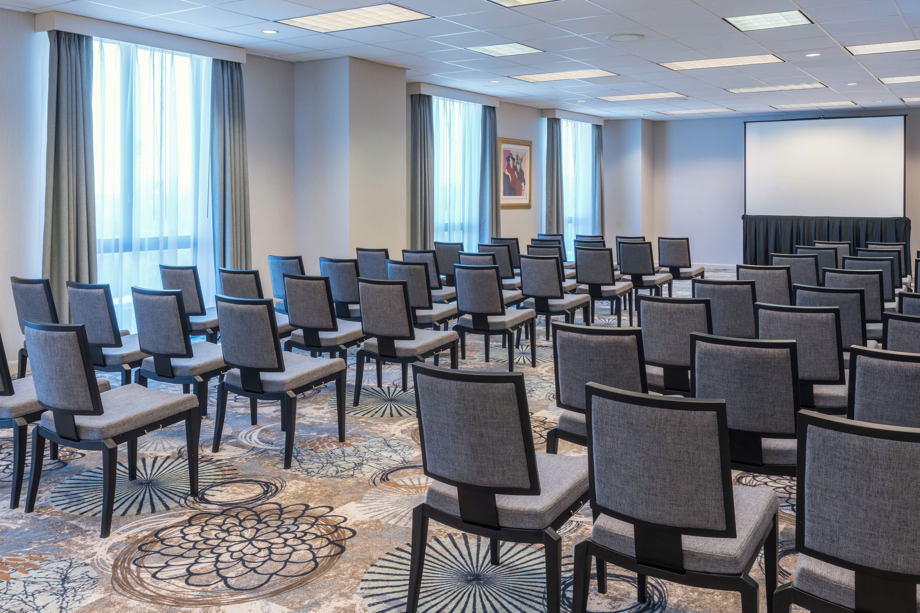 Un salón de conferencias con filas de sillas, ventanas amplias y suelo alfombrado.