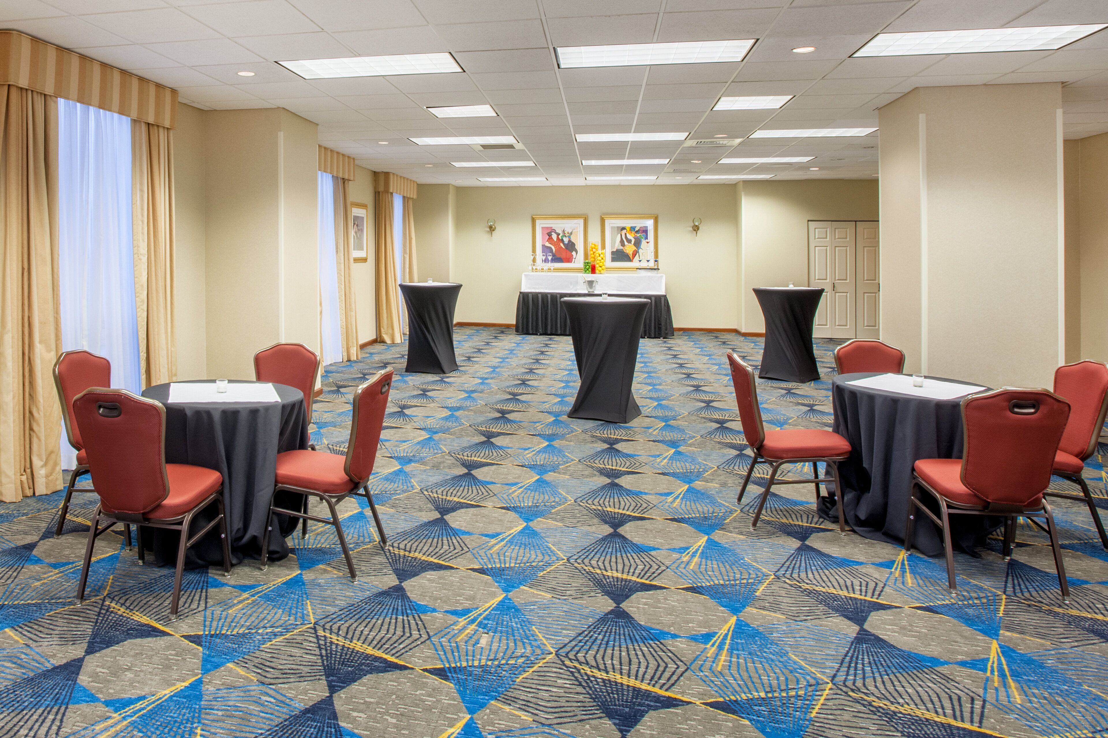 Un salón de conferencias con alfombra de diseño geométrico, mesas redondas con manteles negros y sillas rojas.