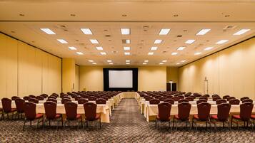 Un salón de conferencias con filas de mesas y sillas, una pantalla de proyector y piso alfombrado.