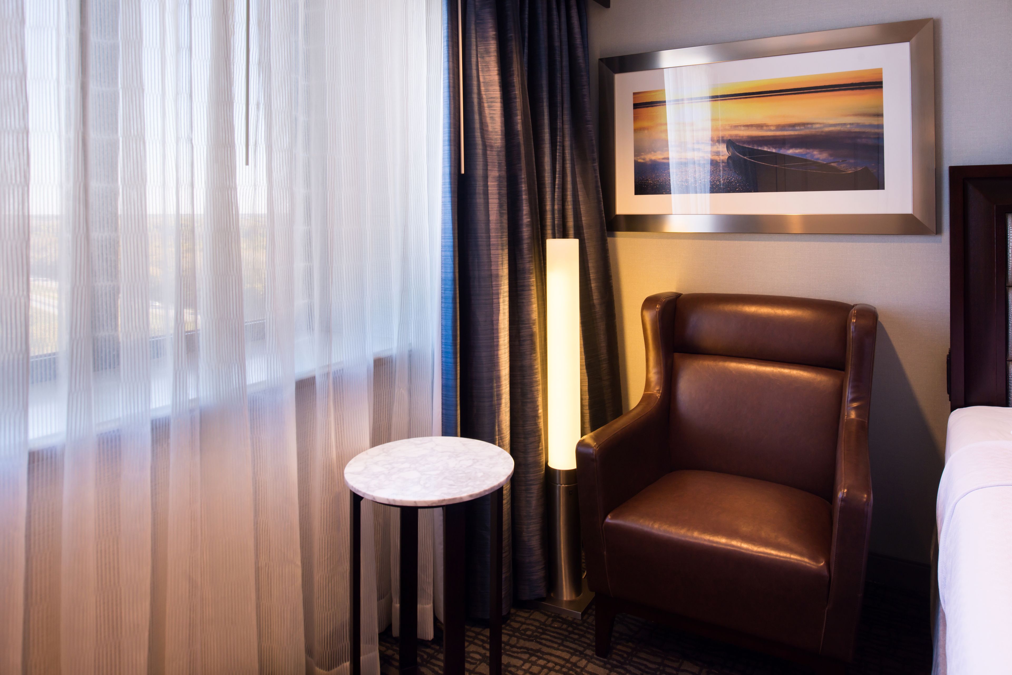 Ein Hotelzimmer mit großem Fenster, braunem Ledersessel, kleinem runden Tisch und einem gerahmten Bild an der Wand.