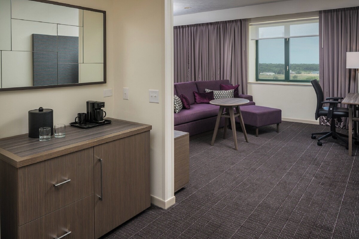 Een hotelkamer met een houten bureau, een paarse bank, een klein rond tafeltje, een stoel en een groot raam met gordijnen.