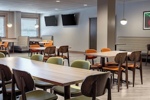 Un espace repas moderne doté de plusieurs tables et chaises, d’un téléviseur à écran plat fixé au mur et d’un coin salon avec un banc.