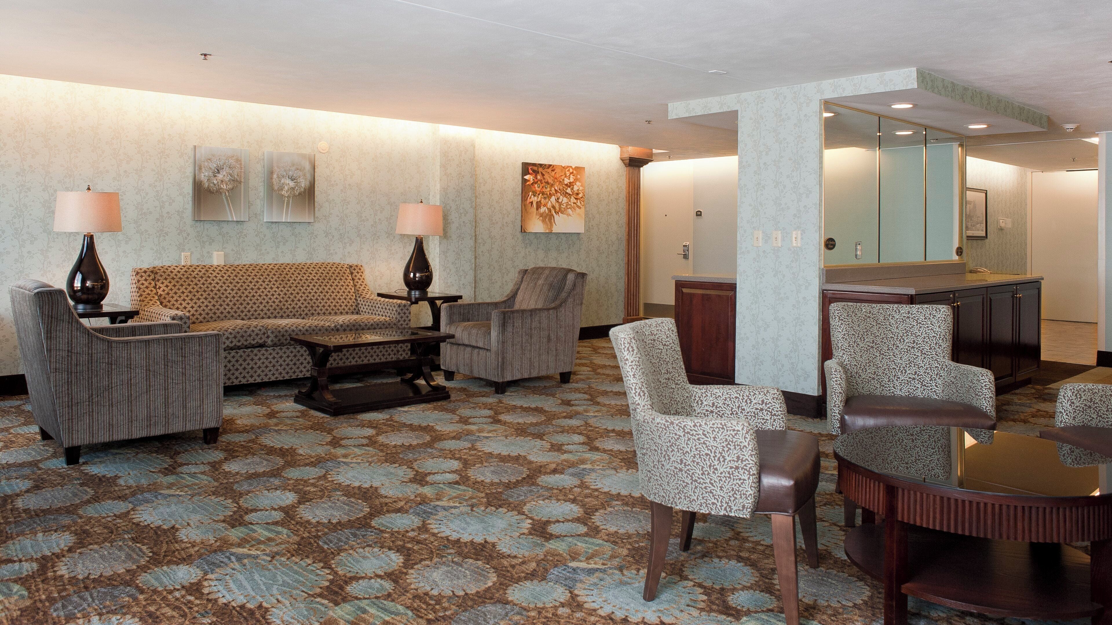 Un vestíbulo de hotel con alfombra estampada, un sofá, sillones, una mesa de centro y un espejo.