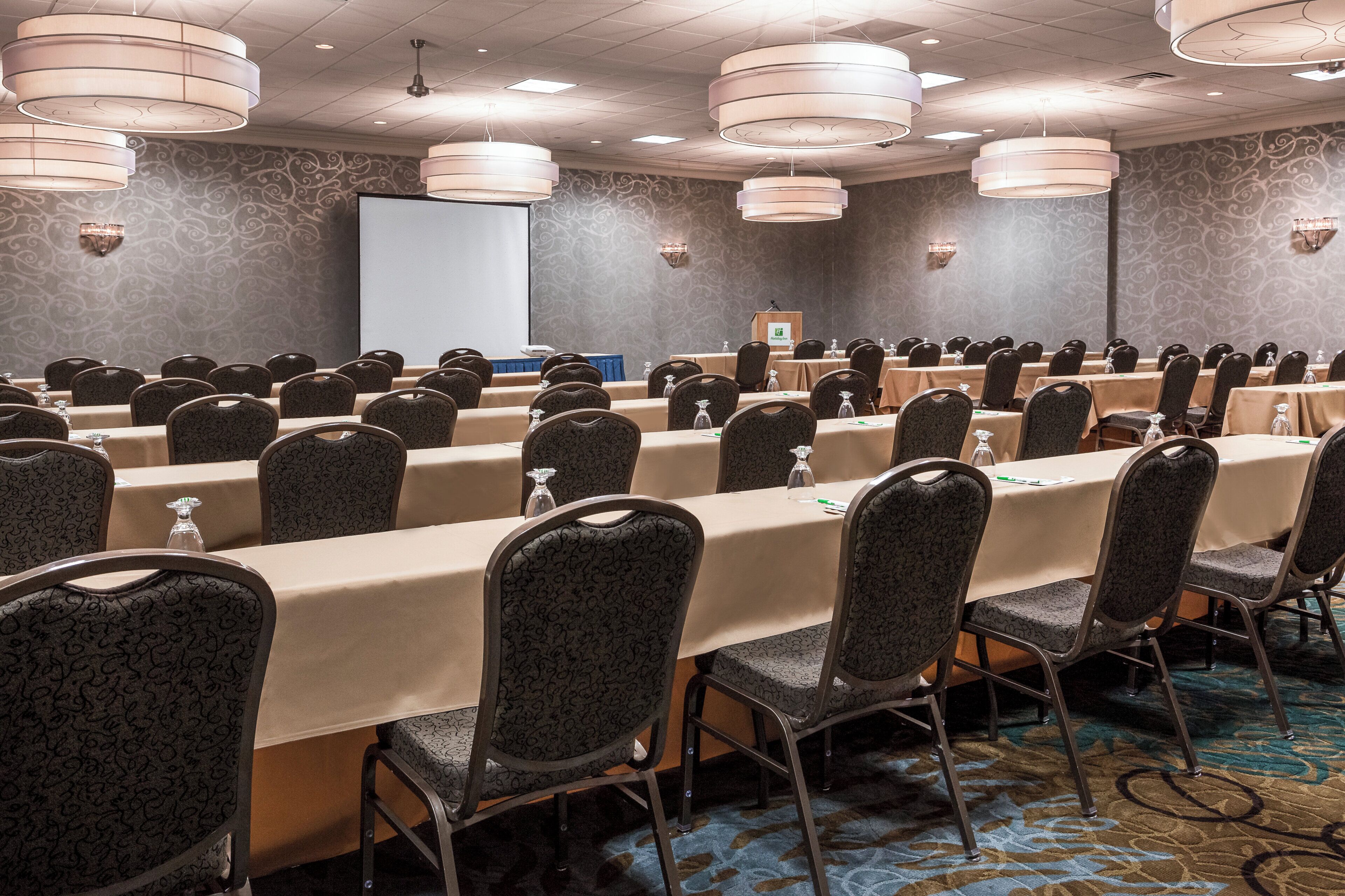 Un salón de conferencias con filas de mesas y sillas, un atril y una pantalla de proyector.