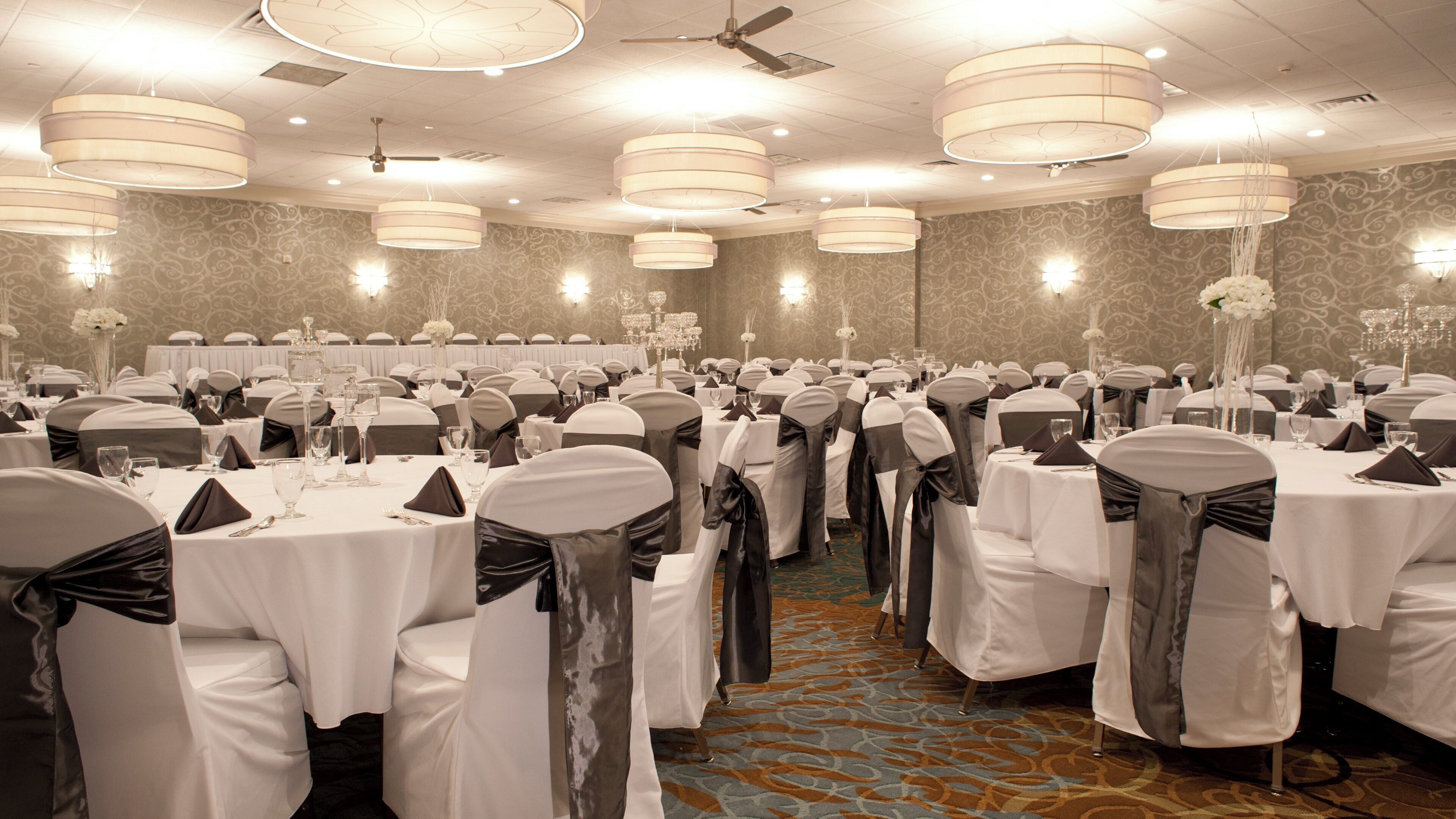 A banquet hall with round tables, chairs with sashes, chandeliers, and a patterned carpet.