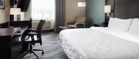 Une chambre d’hôtel avec un grand lit, un bureau avec une chaise, une télévision, une lampe et une fenêtre avec des rideaux.