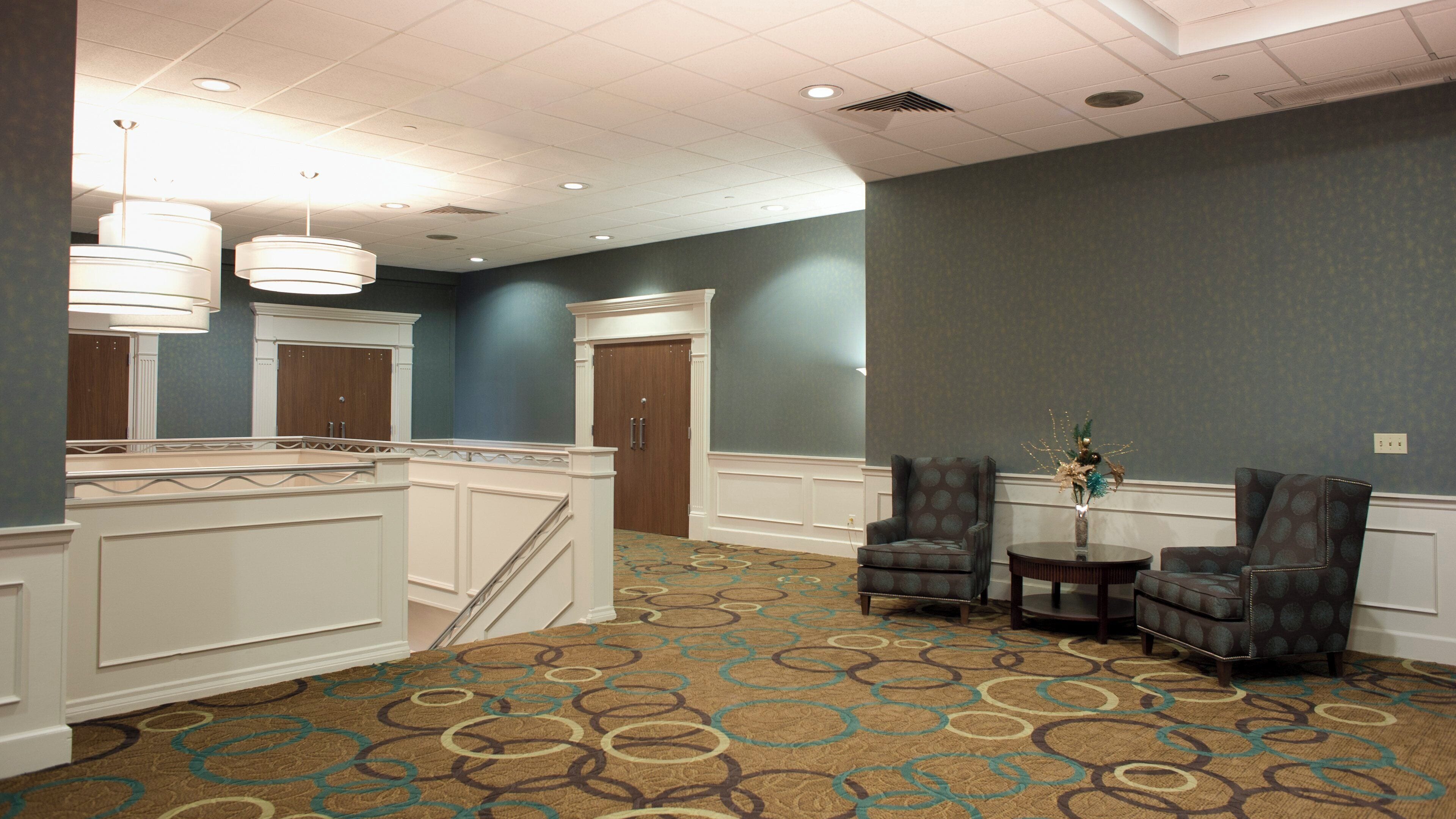 A hotel lobby with a high ceiling, a staircase, two armchairs, a small table with a vase, and a patterned carpet.