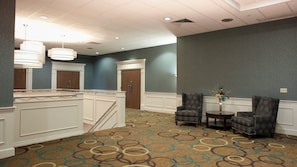 A hotel lobby with a high ceiling, a staircase, two armchairs, a small table with a vase, and a patterned carpet.