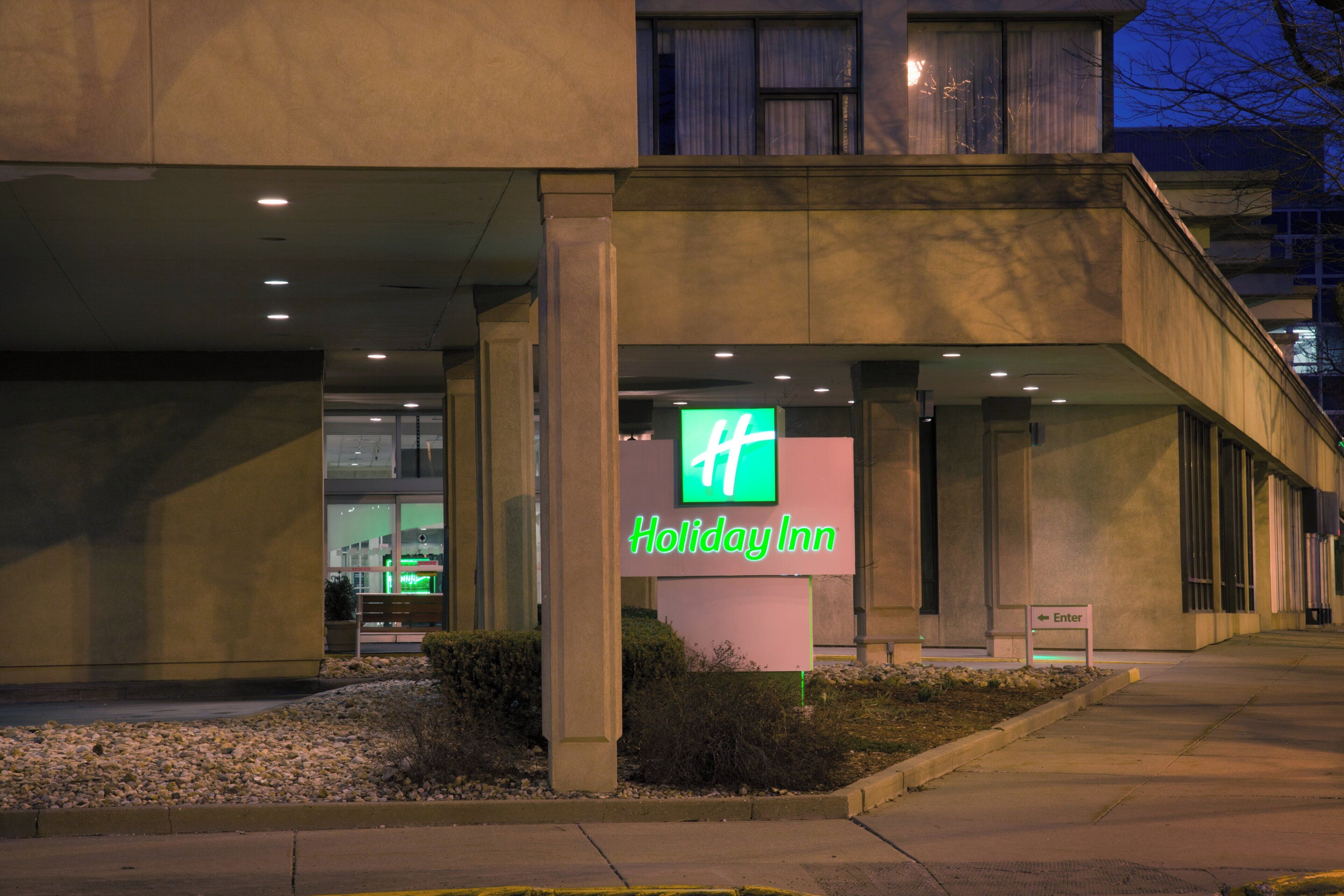 A Holiday Inn hotel entrance with a prominent green sign, a covered walkway, and a landscaped area with rocks and shrubs.