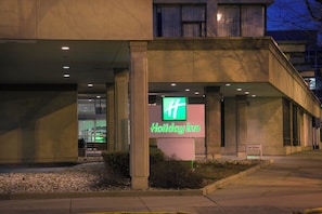A Holiday Inn hotel entrance with a prominent green sign, a covered walkway, and a landscaped area with rocks and shrubs.