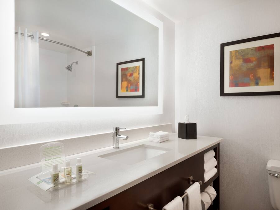 A modern bathroom with a large mirror, a sink, a shower, and a toilet. There are towels and toiletries on the counter.