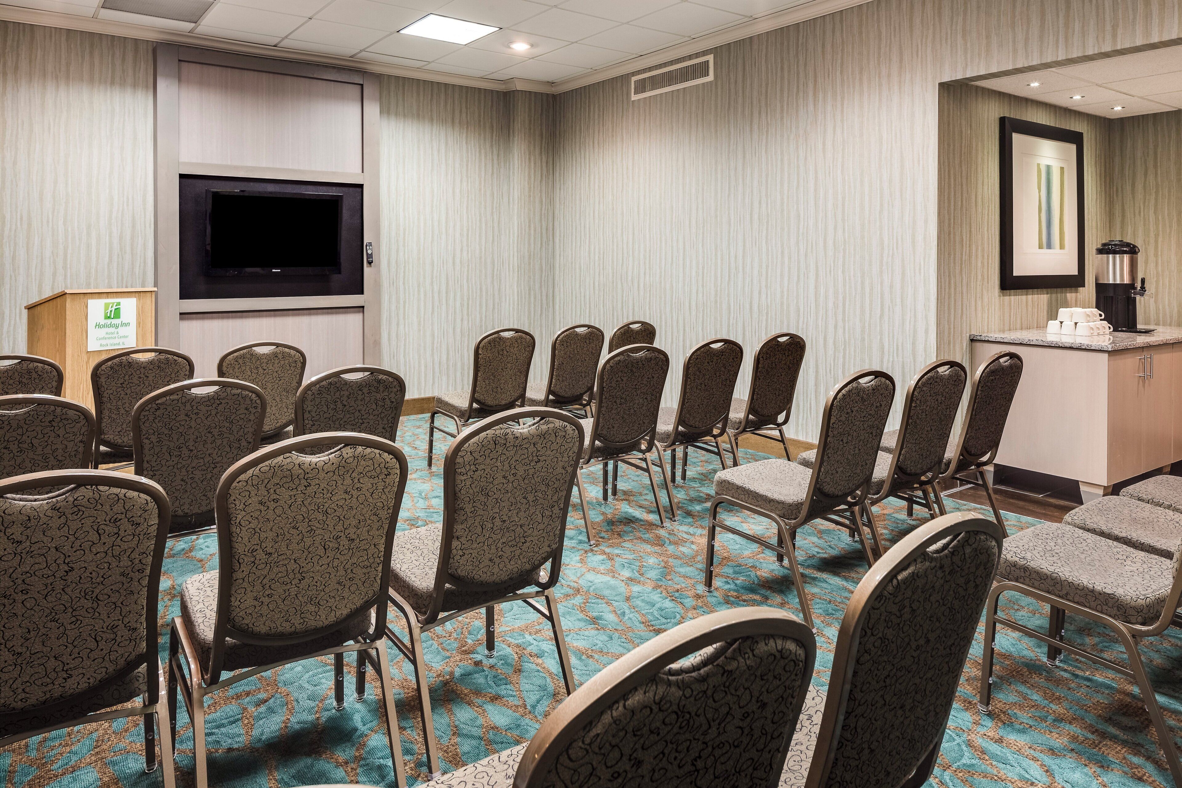 A conference room with rows of chairs, a podium, a flat-screen TV, and a coffee station.