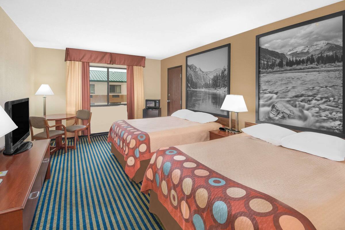 Een hotelkamer met twee bedden, een bureau met een televisie, een stoel, een raam met gordijnen en een groot gepresenteerd beeld van een berglandschap.