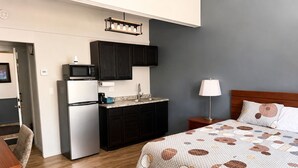 Une chambre d’hôtel compacte avec une kitchenette, un lit recouvert d’une courtepointe à motifs, une lampe de chevet et une tête de lit en bois.