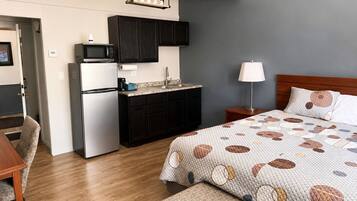 A compact hotel room with a kitchenette, a bed with a patterned quilt, a bedside lamp, and a wooden headboard.