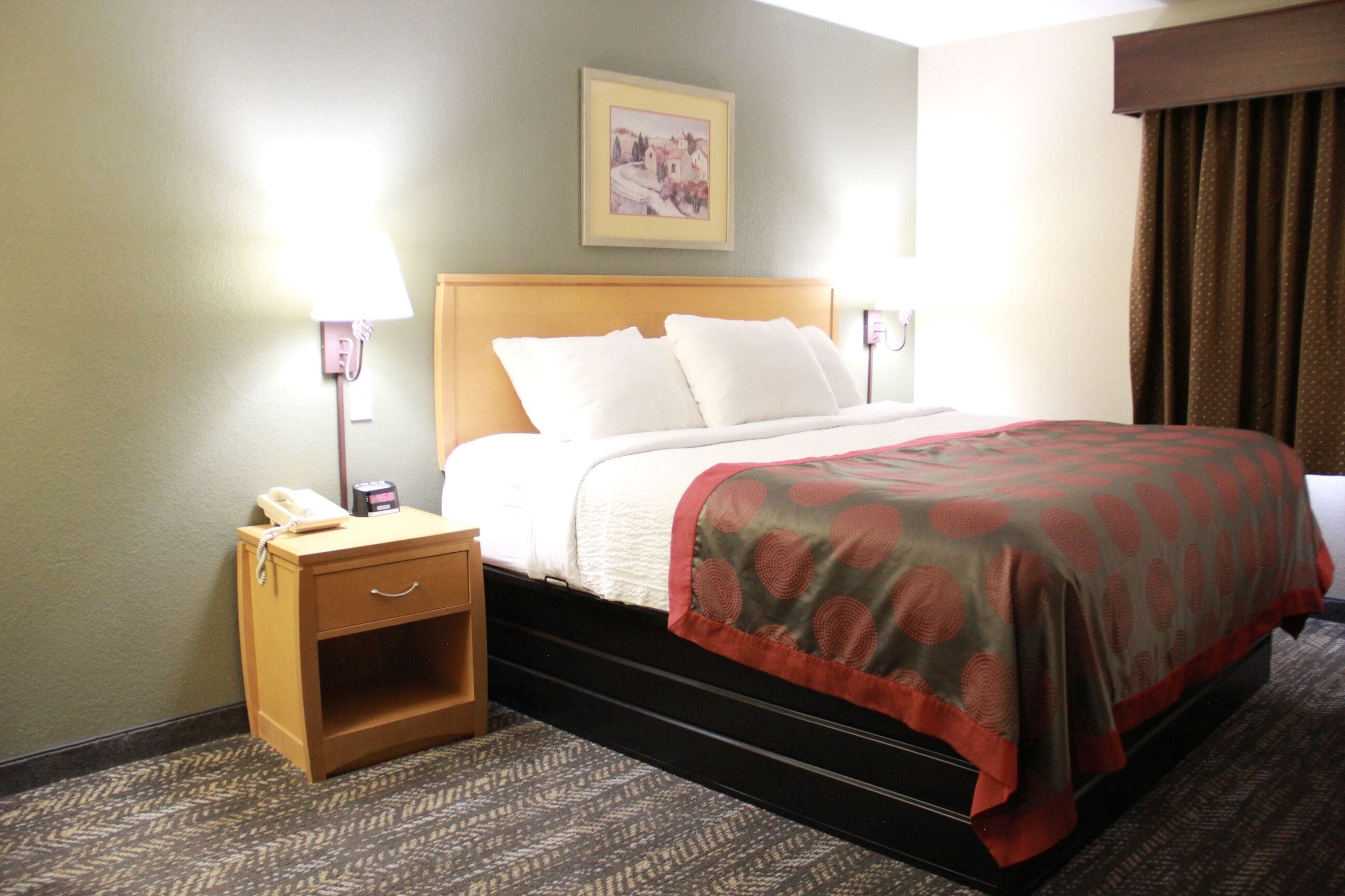 A hotel room with a bed, a nightstand with a phone, a lamp, and a framed picture on the wall.