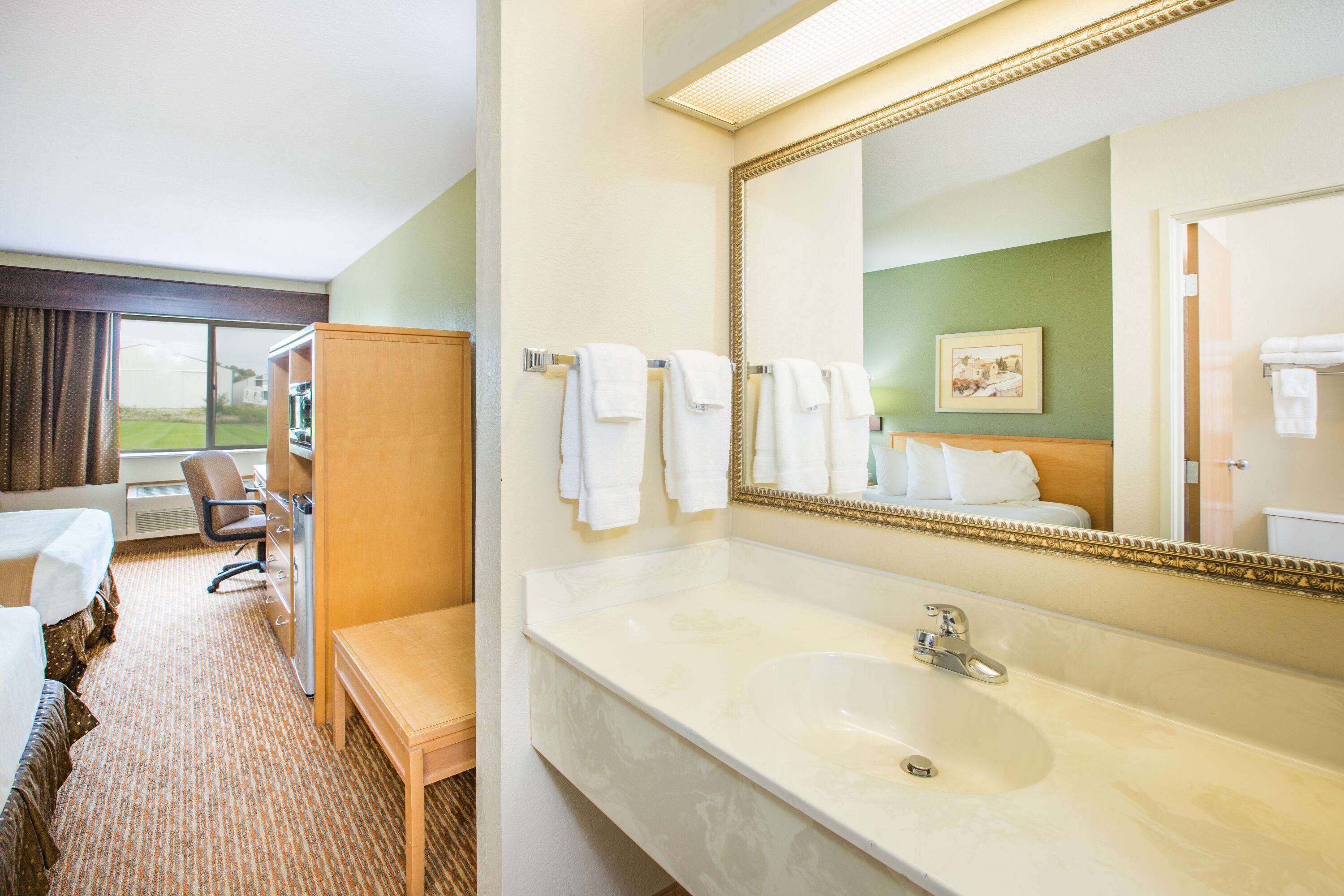 Baño de hotel con un espejo grande, un lavamanos y un banco. Se ve una silla y una cama en la habitación.