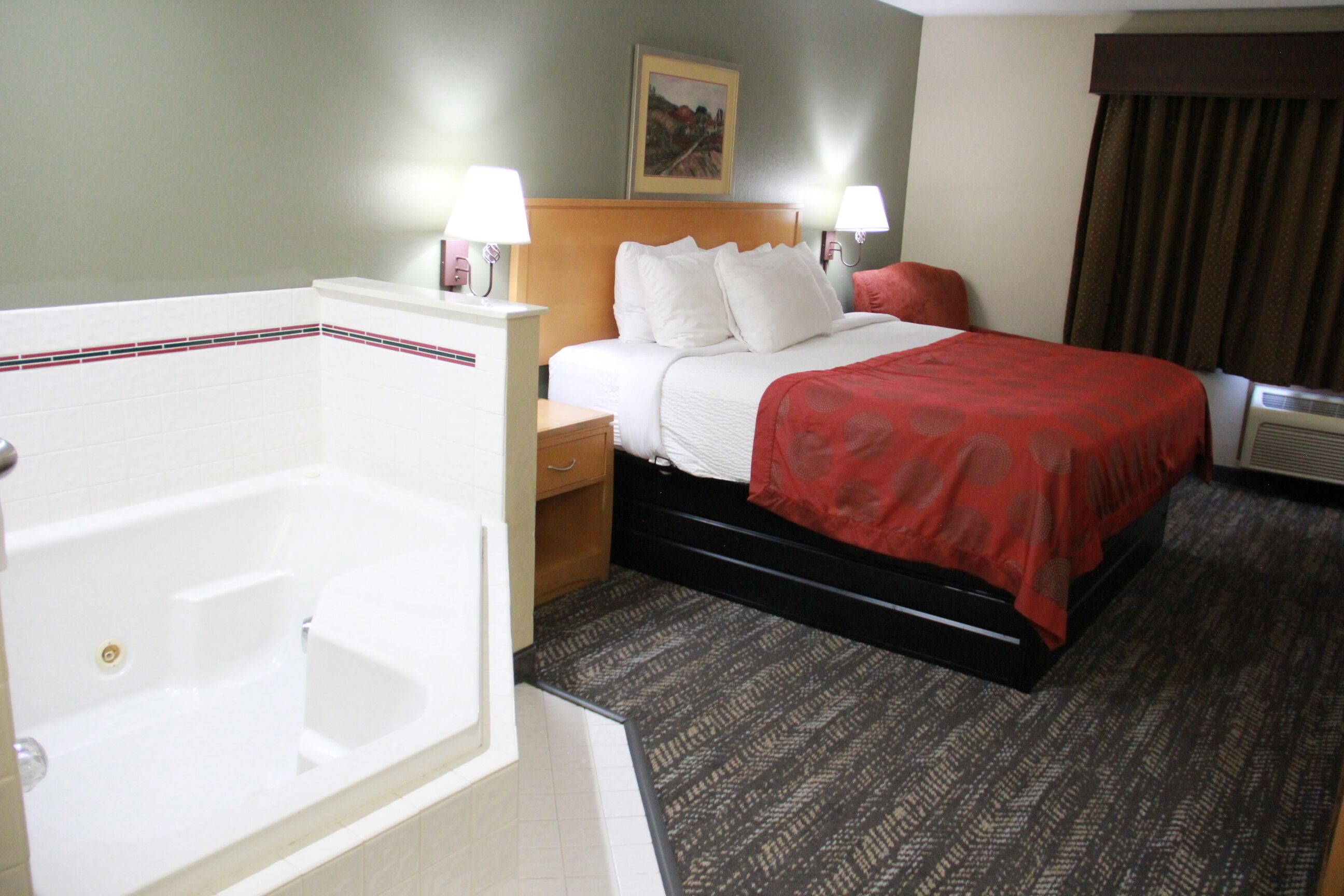A hotel room with a large bathtub, a bed with a red blanket, two bedside lamps, and a painting on the wall.