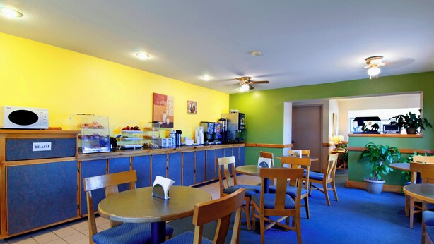 Un restaurante muy iluminado con paredes amarillas, alfombra azul y muebles de madera. Hay mesas y sillas para comer, un bote de basura y un ventilador de techo.
