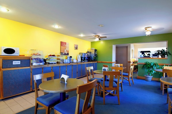 A brightly lit restaurant with yellow walls, blue carpet, and wooden furniture. There are tables and chairs for dining, a trash bin, and a ceiling fan. - Americas Best Value Inn Garden City (Garden City)