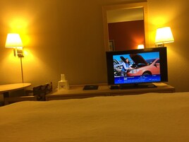 Habitación de hotel con cama, televisor sobre un mueble, dos lámparas de pared y una mesita con una botella y un control remoto.