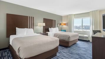 Ein Hotelzimmer mit zwei Betten, einem Schreibtisch, einem Sessel, einem Fernseher und einem großen Fenster mit Vorhängen.