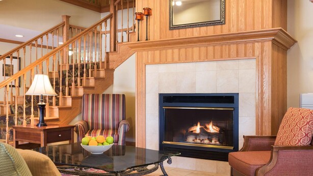 A living room with a fireplace, a glass coffee table with fruit, a striped sofa, and a wooden staircase. - Country Inn & Suites by Radisson, Davenport - Quad Cities (Davenport)
