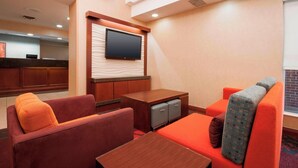 Habitación de hotel con una televisión de pantalla plana montada en la pared, un escritorio de madera, un sofá con cojines naranjas y una mesita.