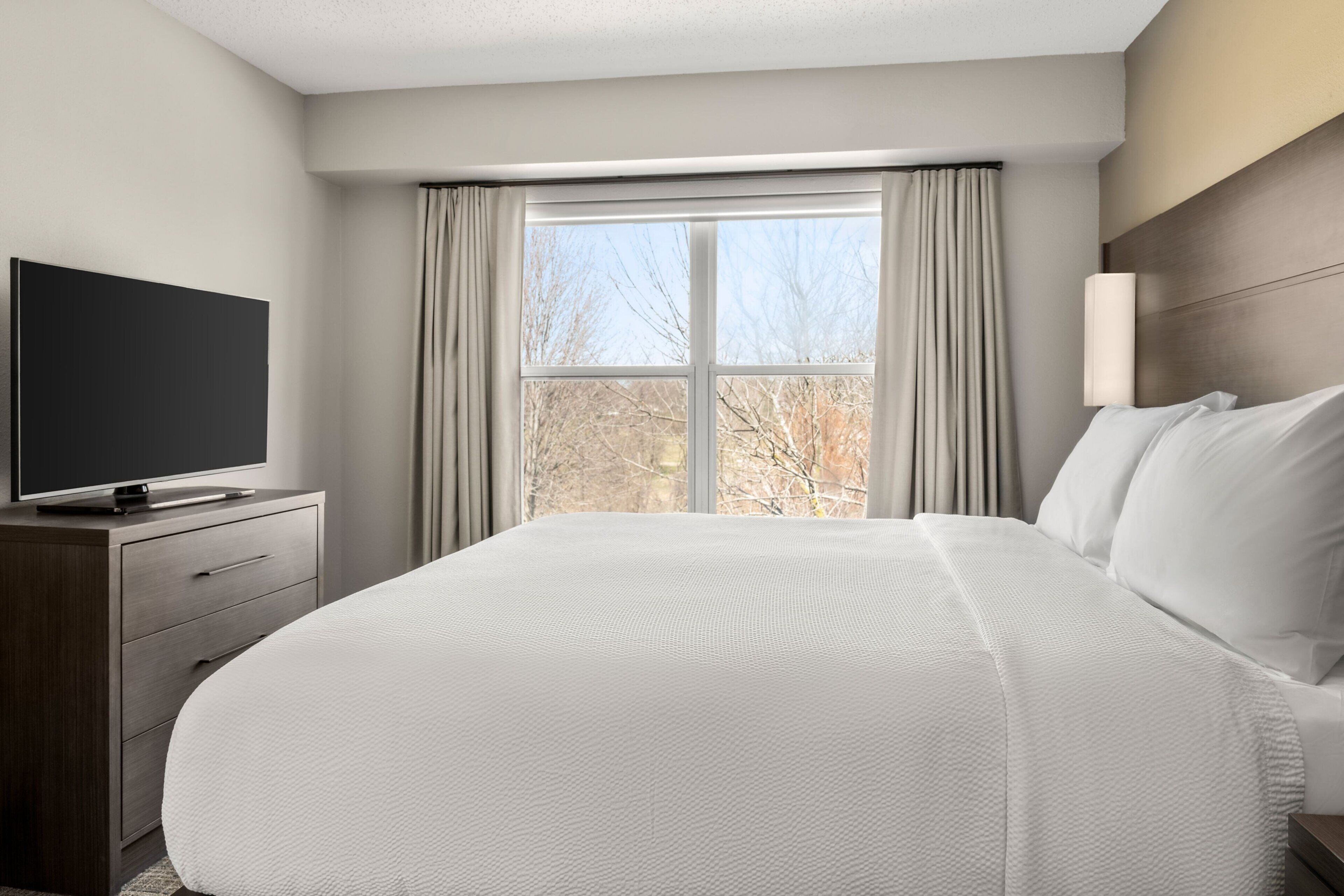 A hotel room with a large bed, a flat-screen TV on a wooden dresser, a window with curtains, and a wall-mounted lamp.
