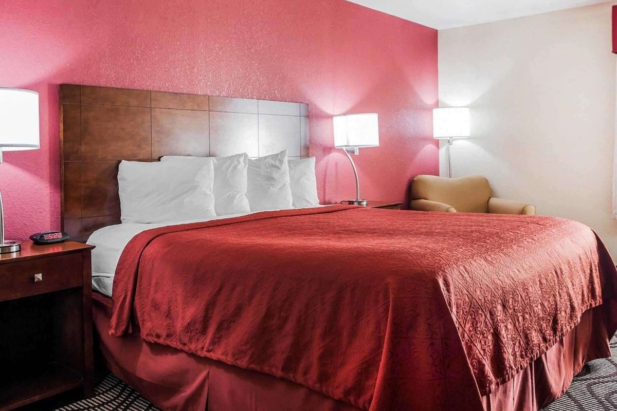 A hotel room with a large bed, two bedside lamps, a wooden headboard, and a red accent wall.