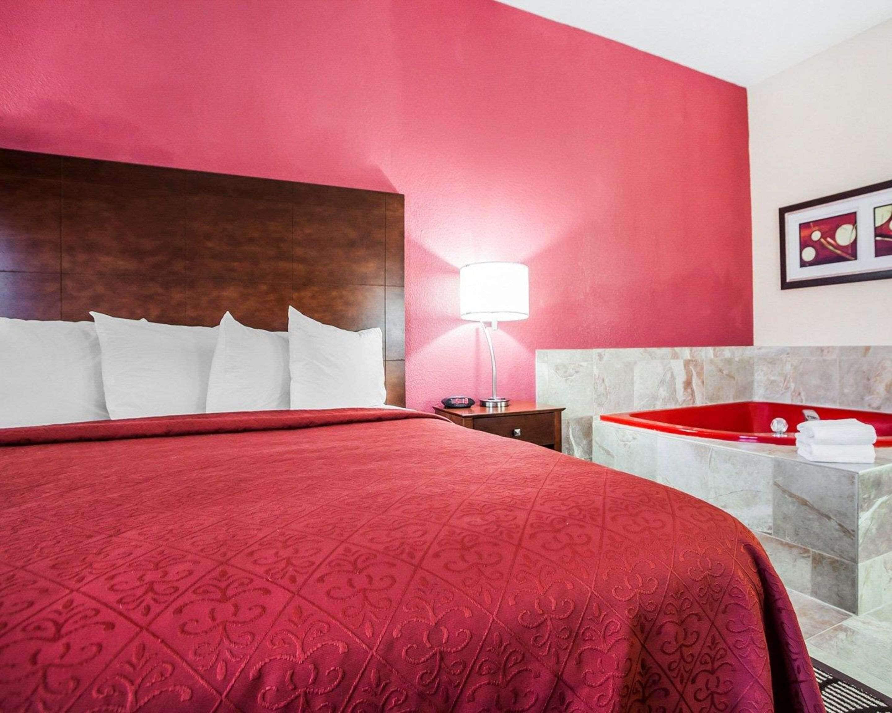 Une chambre d’hôtel avec un grand lit, un mur rouge, une baignoire en marbre et une table de chevet avec une lampe.