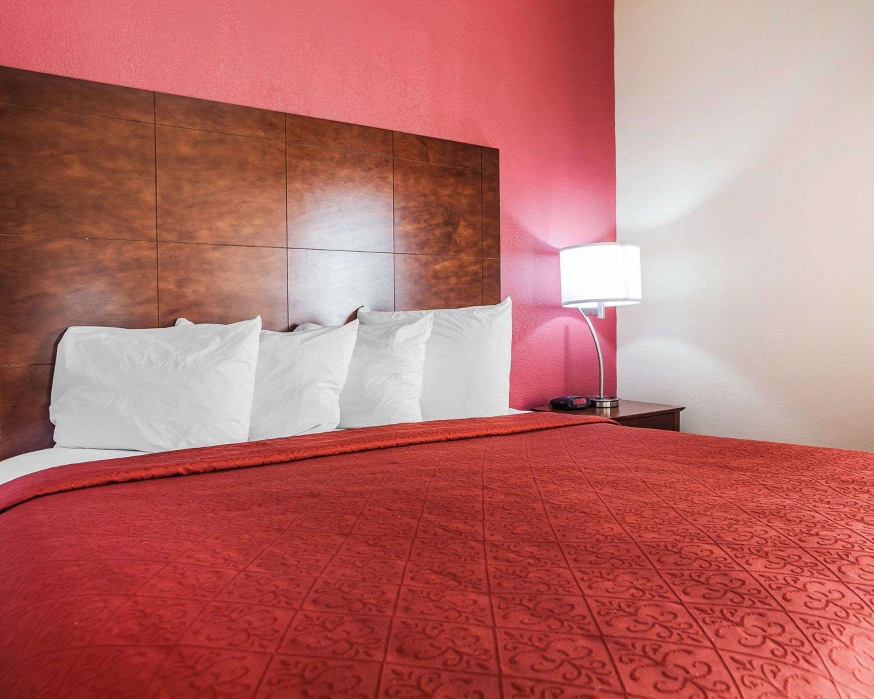 Habitación de hotel con una cama grande, cabecera de madera, colcha con estampado rojo, almohadas blancas, mesita de noche con lámpara y pared roja.