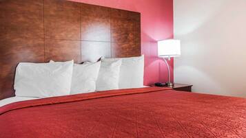 Habitación de hotel con una cama grande, cabecera de madera, colcha con estampado rojo, almohadas blancas, mesita de noche con lámpara y pared roja.