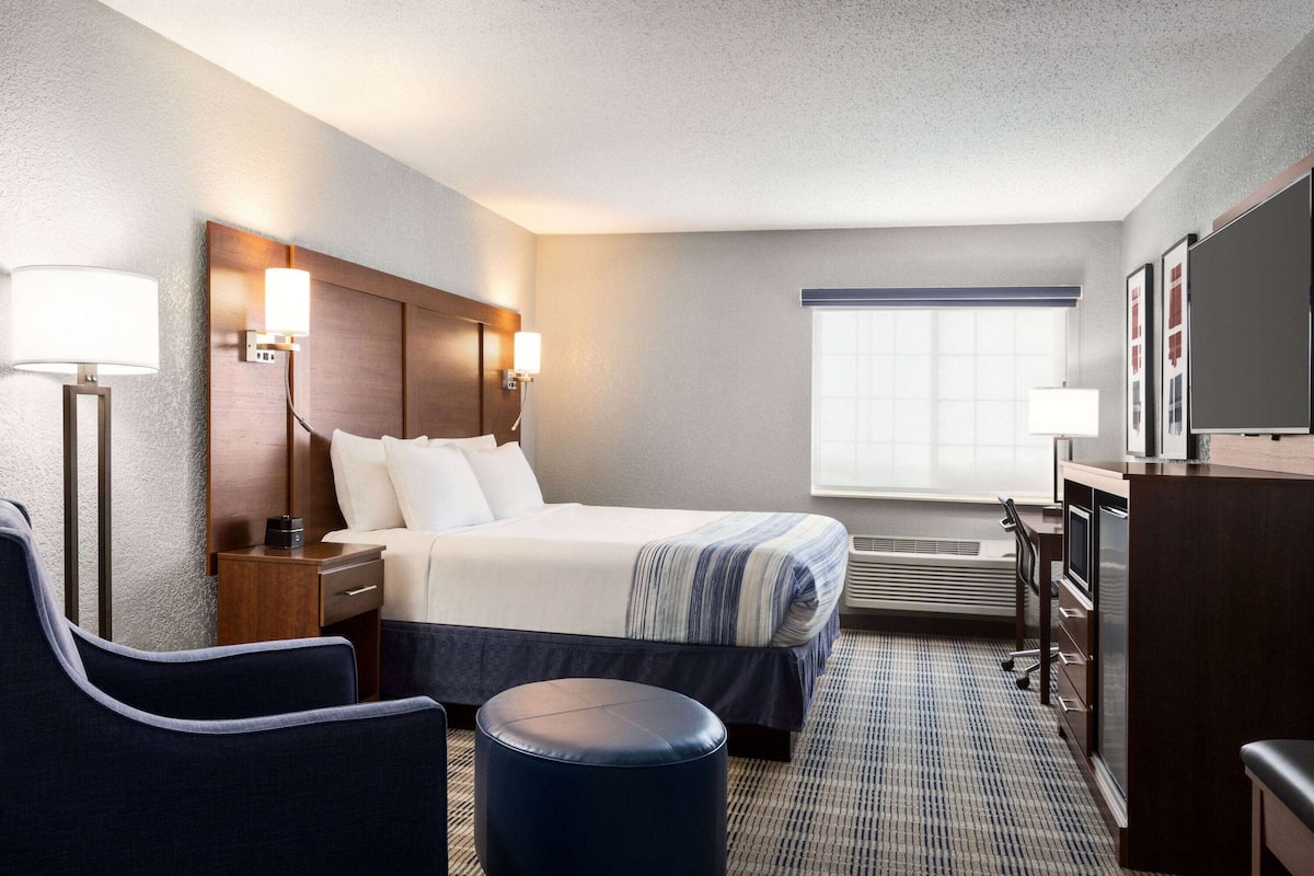 A hotel room with a bed, bedside tables, a desk, a chair, a round ottoman, and a large window.