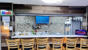 A bar area with wooden chairs, a neon sign, a digital display, and a refrigerator. - AmericInn by Wyndham Williamsburg (South Amana)