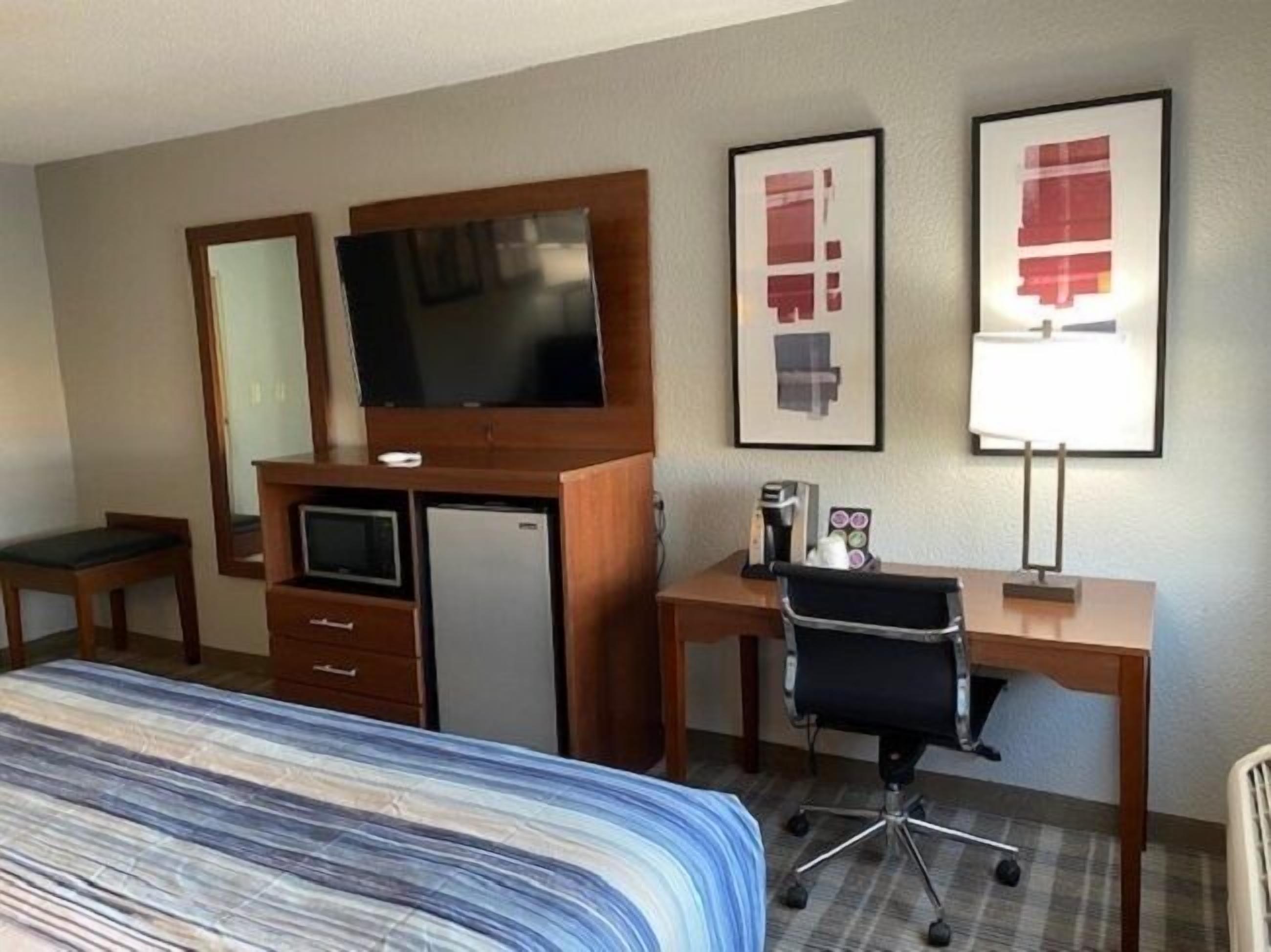 A hotel room with a bed, a desk with a chair, a television, a refrigerator, and artwork on the wall.