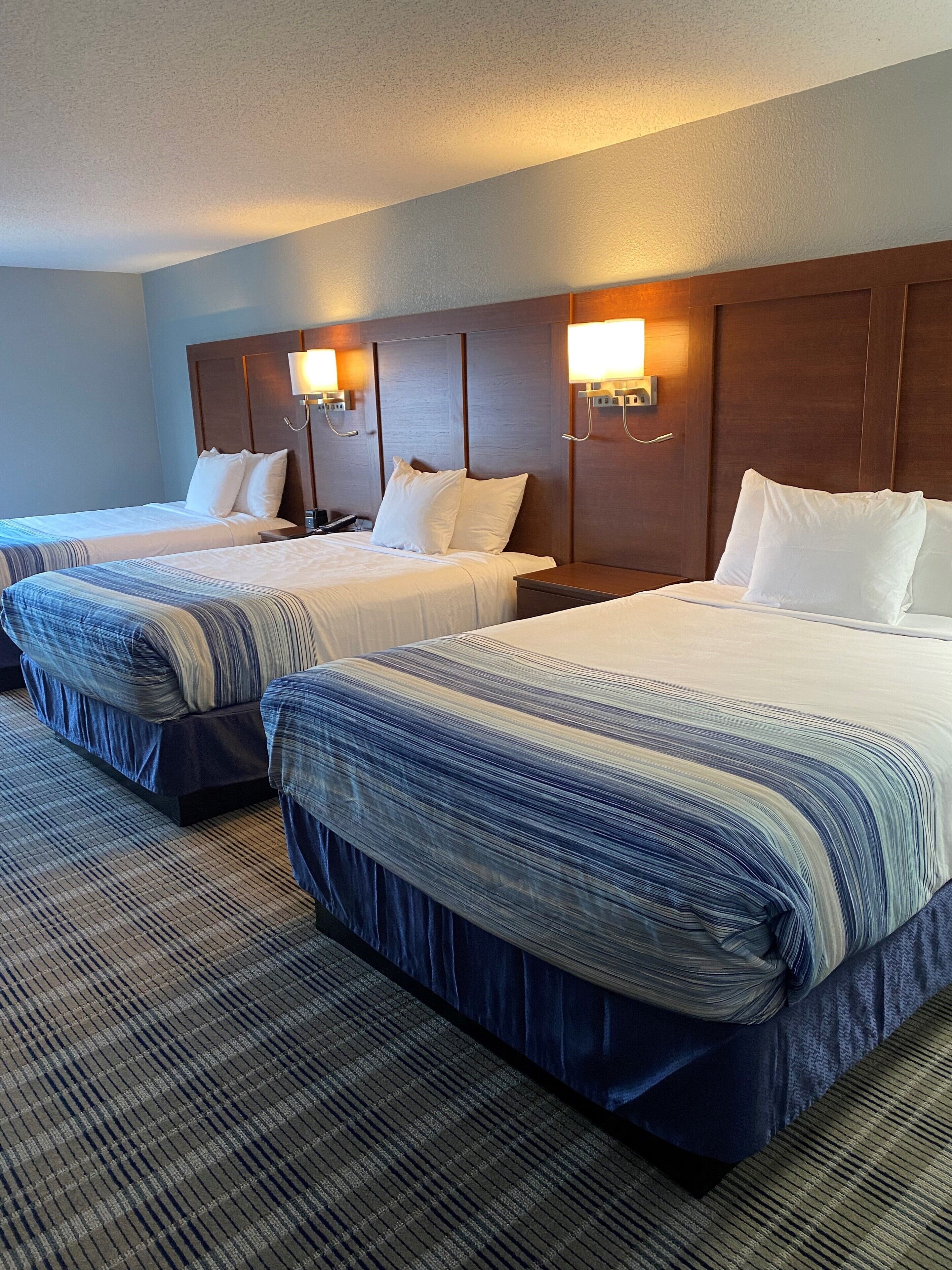 Quarto de hotel com duas camas, cada uma com colcha listrada, cabeceira de madeira e luminárias fixadas na parede.