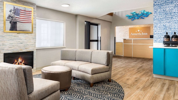 A modern hotel lobby with a sectional sofa, a fireplace, a reception desk, and a decorative wall with an American flag. - AmericInn by Wyndham Williamsburg (South Amana)